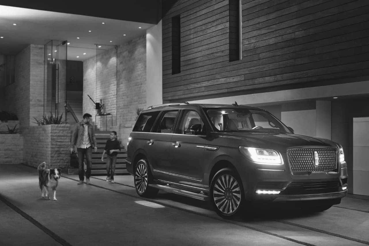 Luxury SUV parked outside modern home, with two people and a dog approaching.