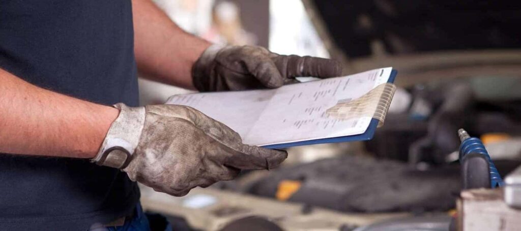 Mechanic in gloves holding a clipboard near an open car hood in a garage setting.