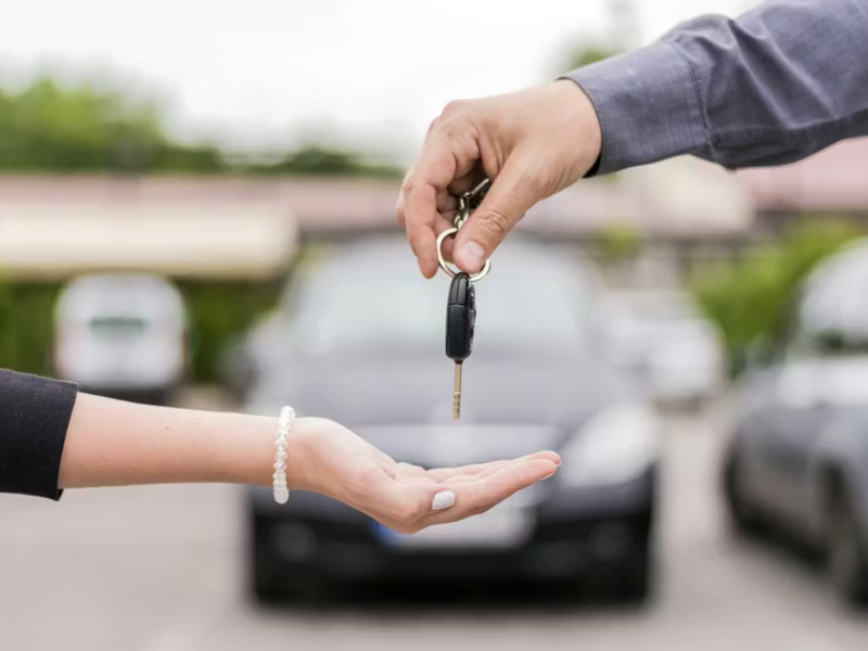 Hand giving car keys to another person, with a car in the background.